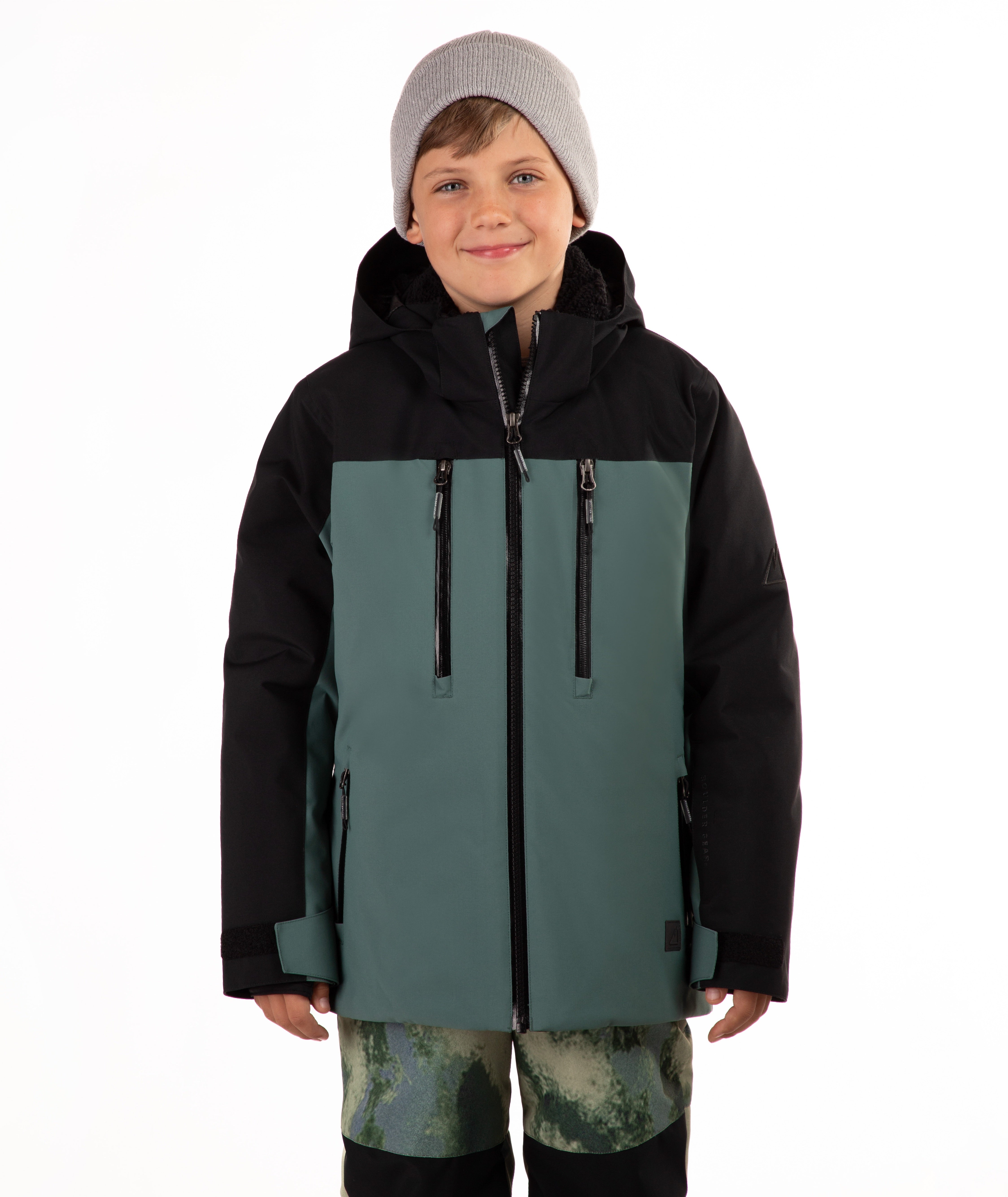 A child wearing a color-blocked ski jacket in black and muted teal, paired with a gray beanie and camouflage-patterned pants, against a plain white background.