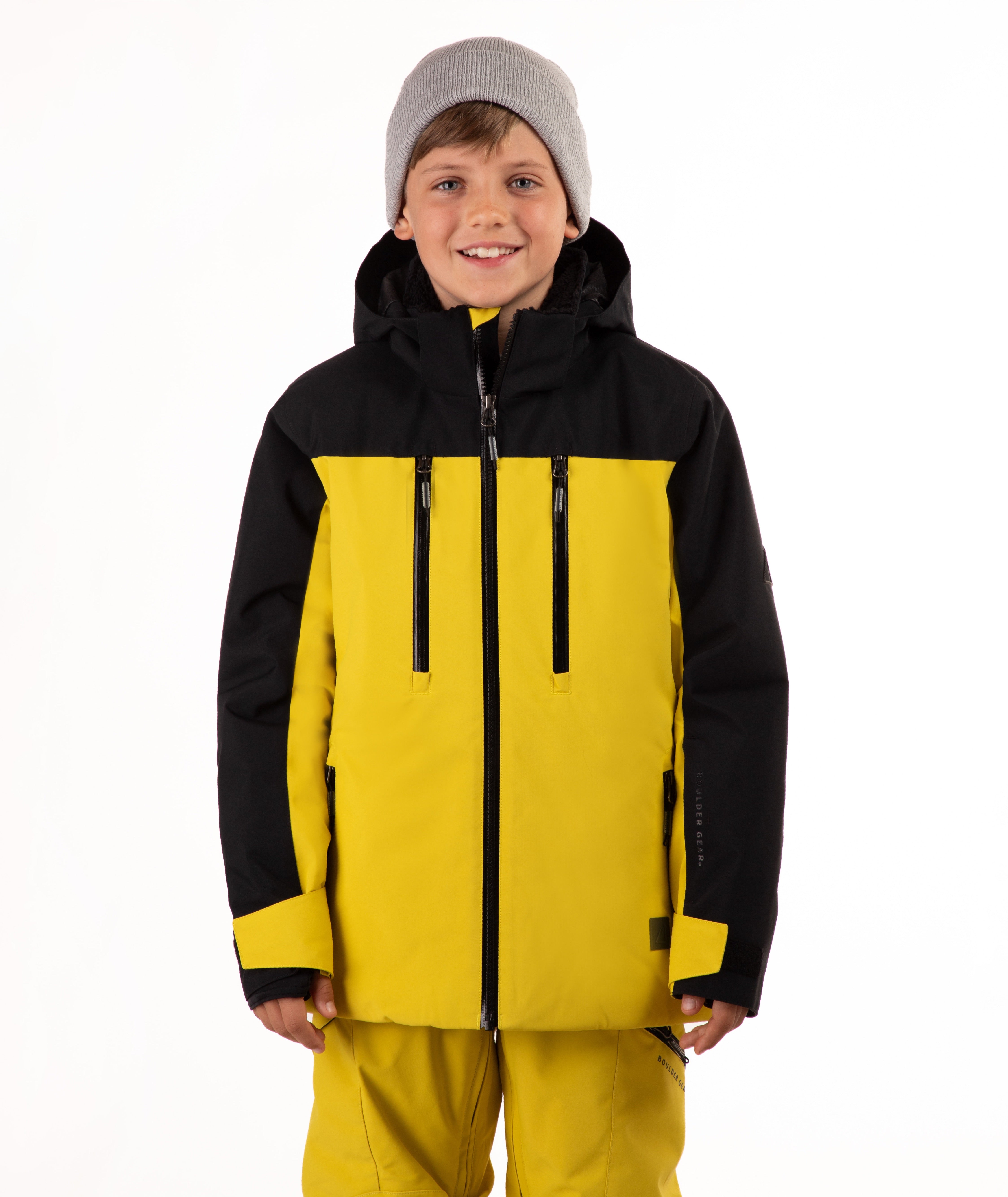 A smiling young boy wearing a bright yellow and black ski jacket with matching yellow ski pants and a gray knit beanie, photographed against a plain white studio background.