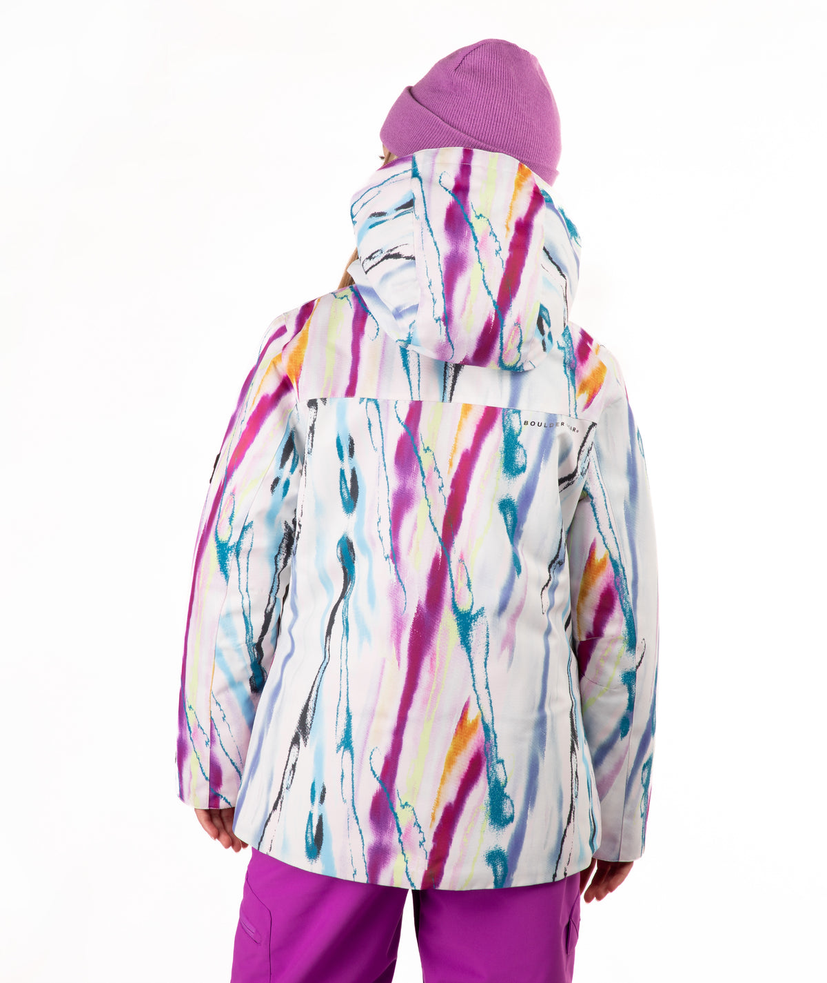 A child viewed from the back wearing a hooded ski jacket with a colorful abstract paint-stroke pattern in pink, purple, blue, yellow, and orange on a white base, paired with a purple beanie and purple pants, against a plain white background.