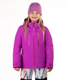 A young girl wearing a bright pin-purple ski jacket, a matching knit beanie, and colorful patterned ski pants, standing against a plain white studio background.