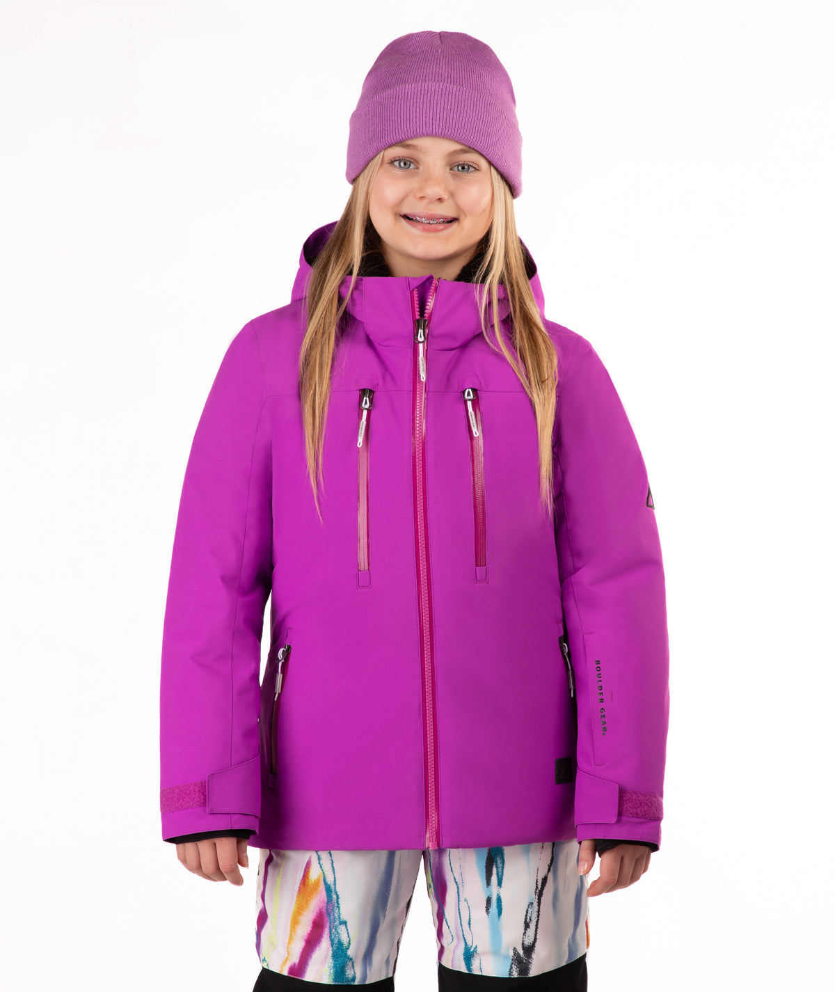 A young girl wearing a bright pin-purple ski jacket, a matching knit beanie, and colorful patterned ski pants, standing against a plain white studio background.