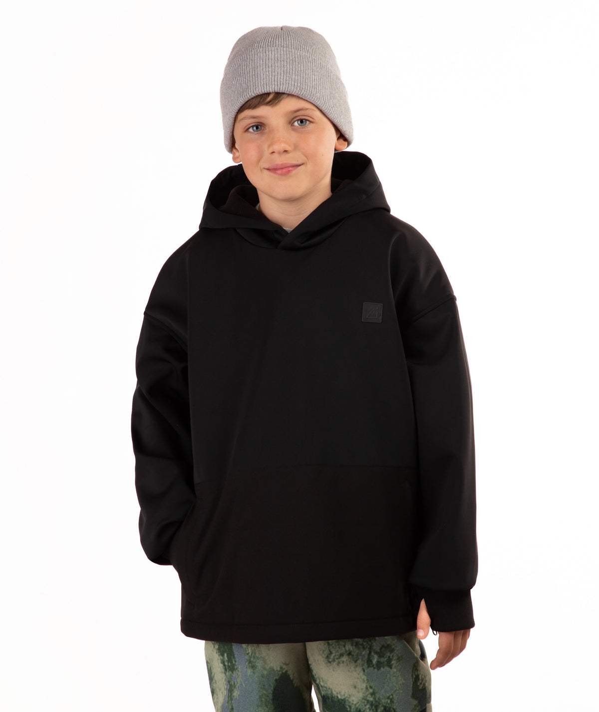 A child wearing a black pullover hoodie with a small tonal square logo patch on the left chest, paired with a light gray knit beanie and camouflage-patterned pants, photographed against a plain white background.