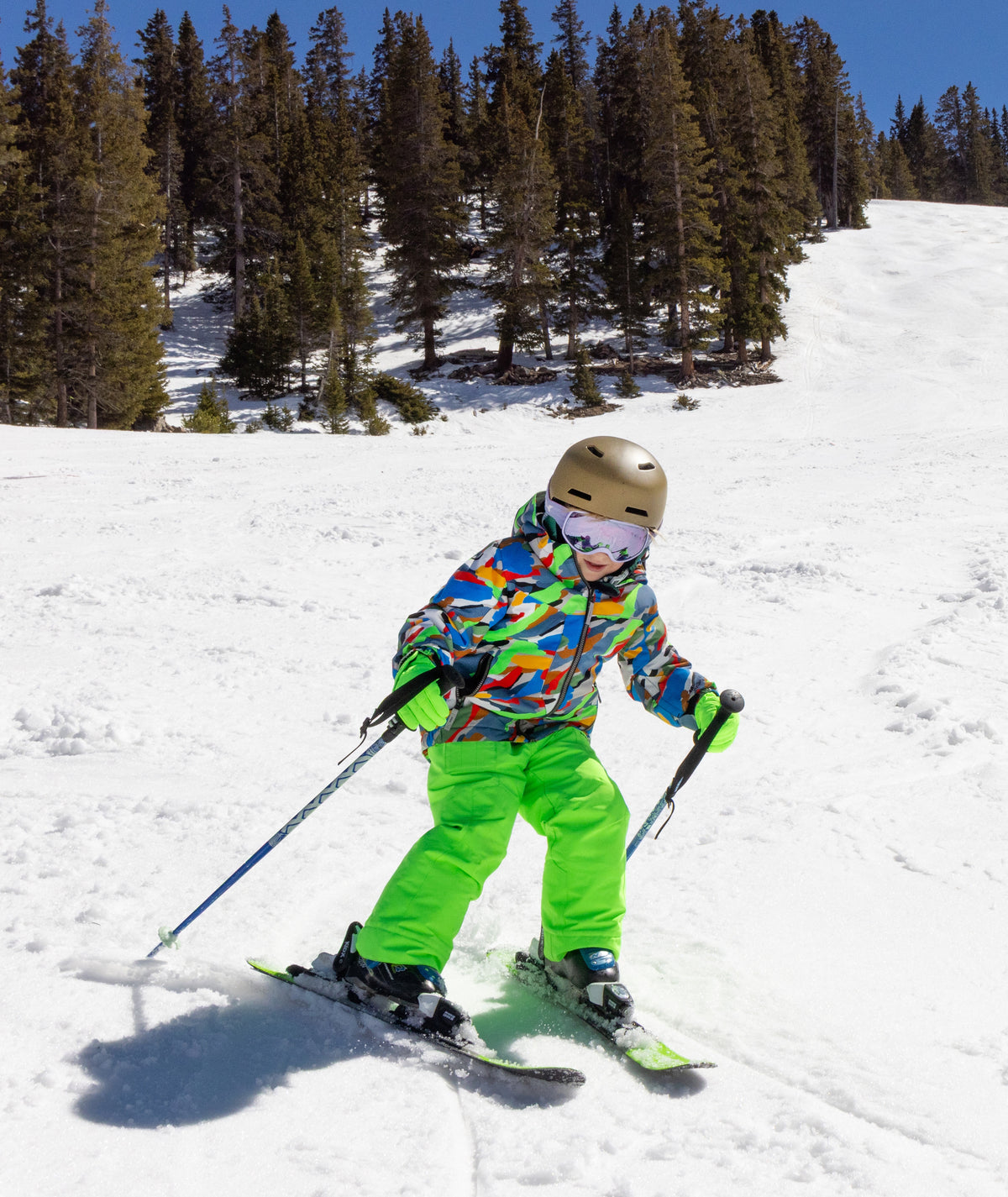 A child skier wearing a colorful patterned jacket, bright green pants, a gold helmet, and goggles is skiing down a snowy mountain slope with pine trees in the background.