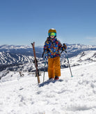 A child skier stands on a snowy mountain slope wearing a colorful patterned ski jacket, bright orange pants, a yellow helmet, and ski goggles, with skis and poles in hand. Snow-covered mountains and a clear blue sky are visible in the background.