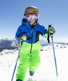 A child skier wearing a bright blue and neon green ski jacket and pants, along with a helmet, goggles, and ski poles, posing on a snowy mountain slope under a clear blue sky.