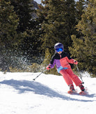 a child skiing down a snowy tree lined path wearing a multi colored black, pink, purple and blue jacket with pink pants.