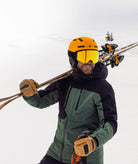 A smiling skier wearing a green and black ski jacket, a bright yellow helmet, and yellow mirrored goggles, carrying ski poles over his shoulder on a snowy mountain.