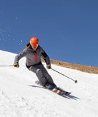 A skier carving through fresh powder on a snowy slope under a clear blue sky, wearing a gray ski jacket, dark pants, a bright orange helmet, and orange-tinted goggles.