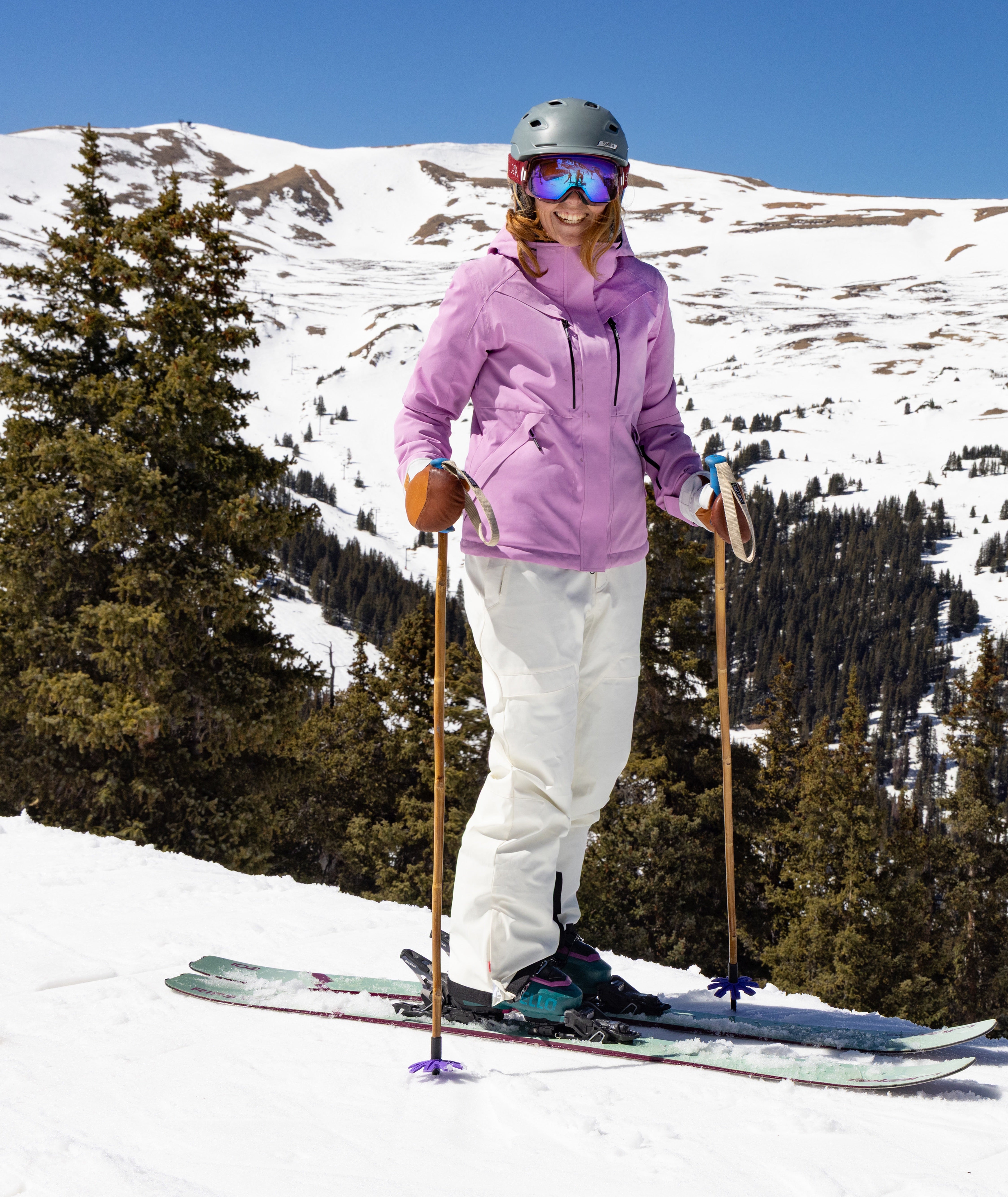 woman skier on a mountain side wearing a light purple jacket, gray helmet and white pants. a snowy mountain and pine trees can be seen in the distance.