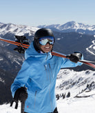 woman shown on a mountain side smiling while wearing a light blue jacket, holding skis. snow covered mountains can be seen in the background.