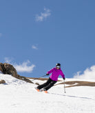 woman shown skiing on a mountain top wearing a bright purple-pink jacket with black pants and helmet.