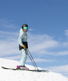 woman shown on a snowy mountain top with clear blue skies wearing a light green and black jack, light green pants.