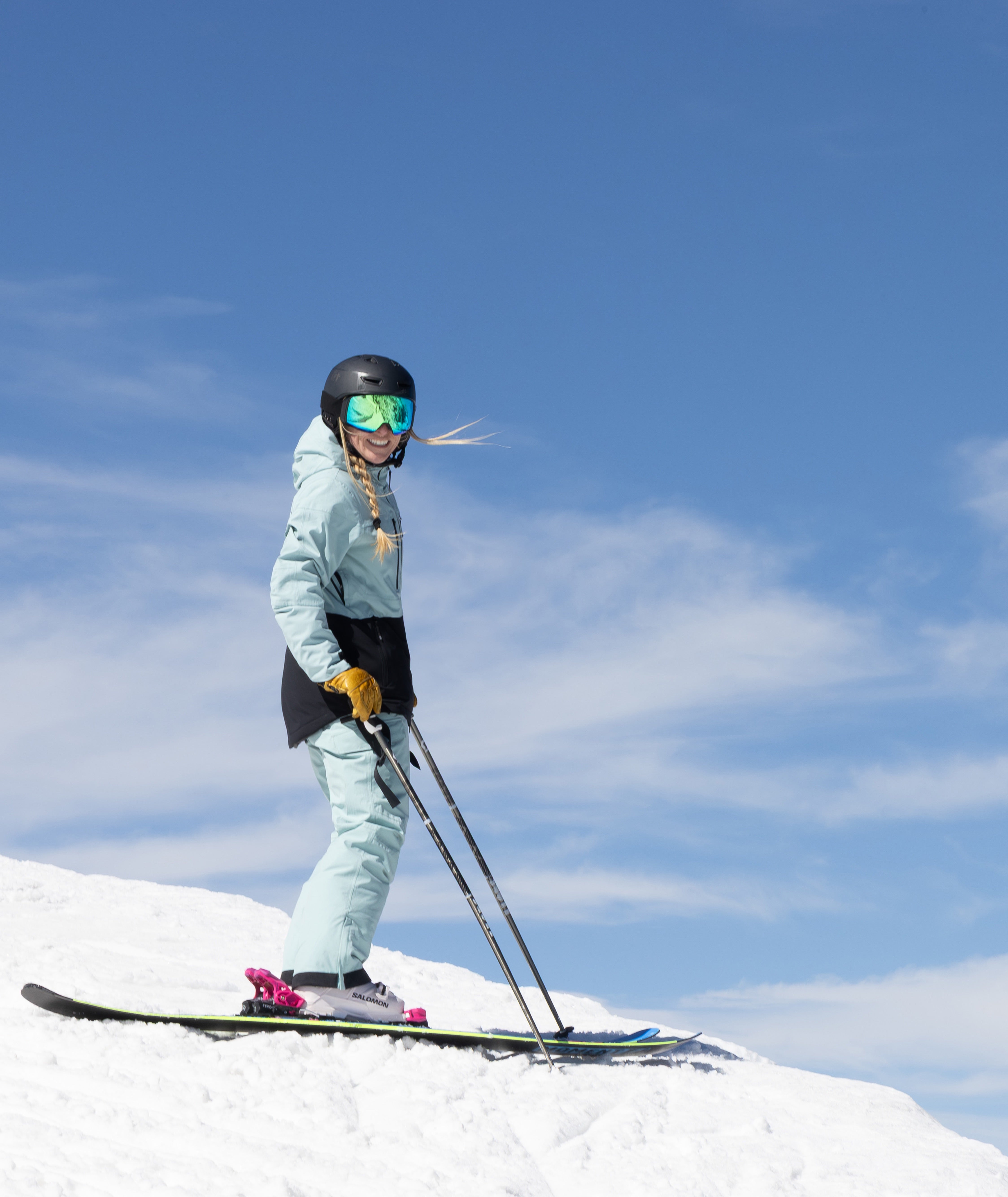 woman shown on a snowy mountain top with clear blue skies wearing a light green and black jack, light green pants.