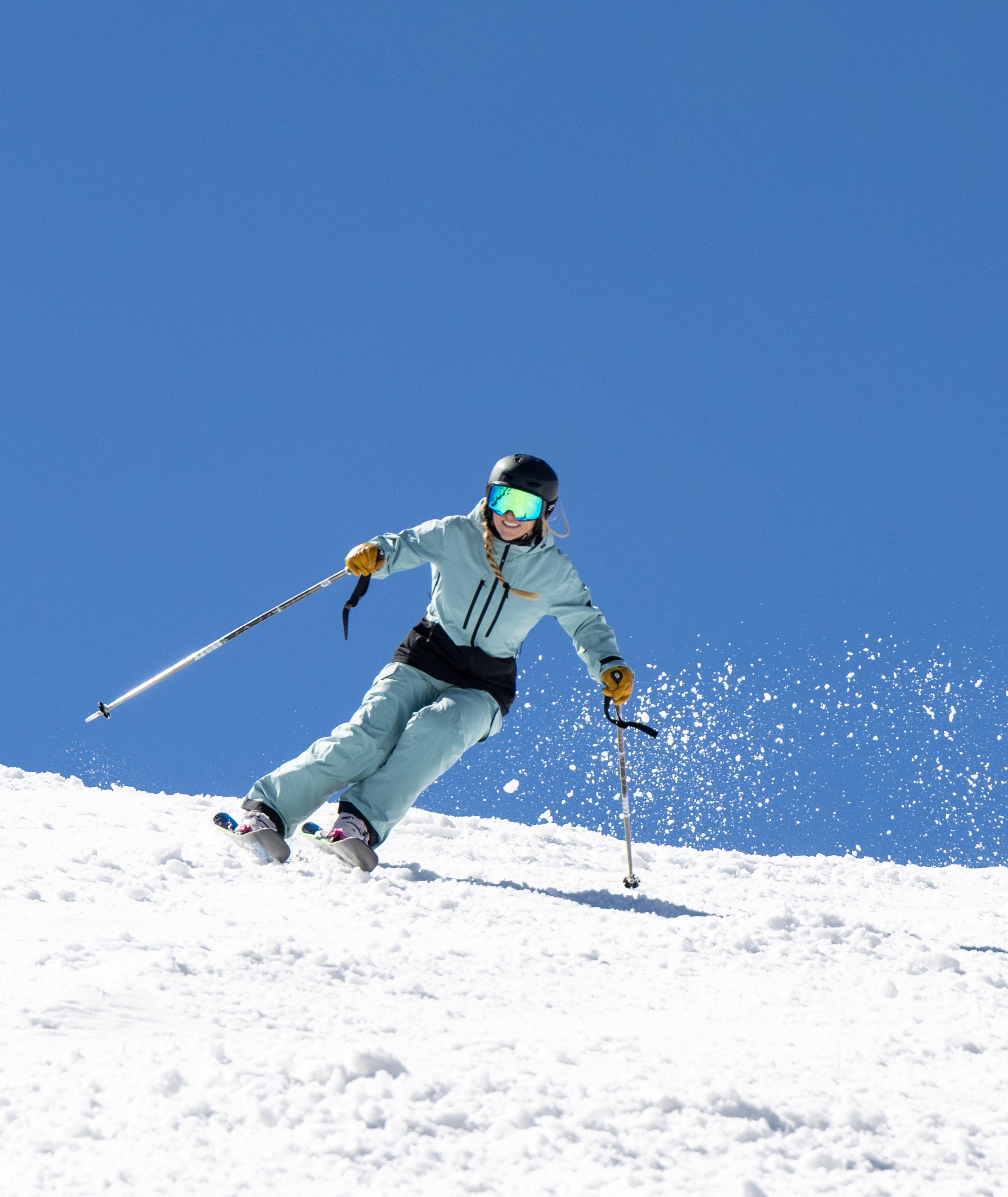 A skier in a light green jacket and matching pants glides through fresh snow on a sunny day, with a bright blue sky overhead and snow spray visible behind them.