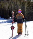 A young person standing in the snow wearing ski gear, including a tan and dark green ski jacket, brown ski pants, an orange helmet, and purple goggles, holding a ski and ski poles with a ski standing vertically.