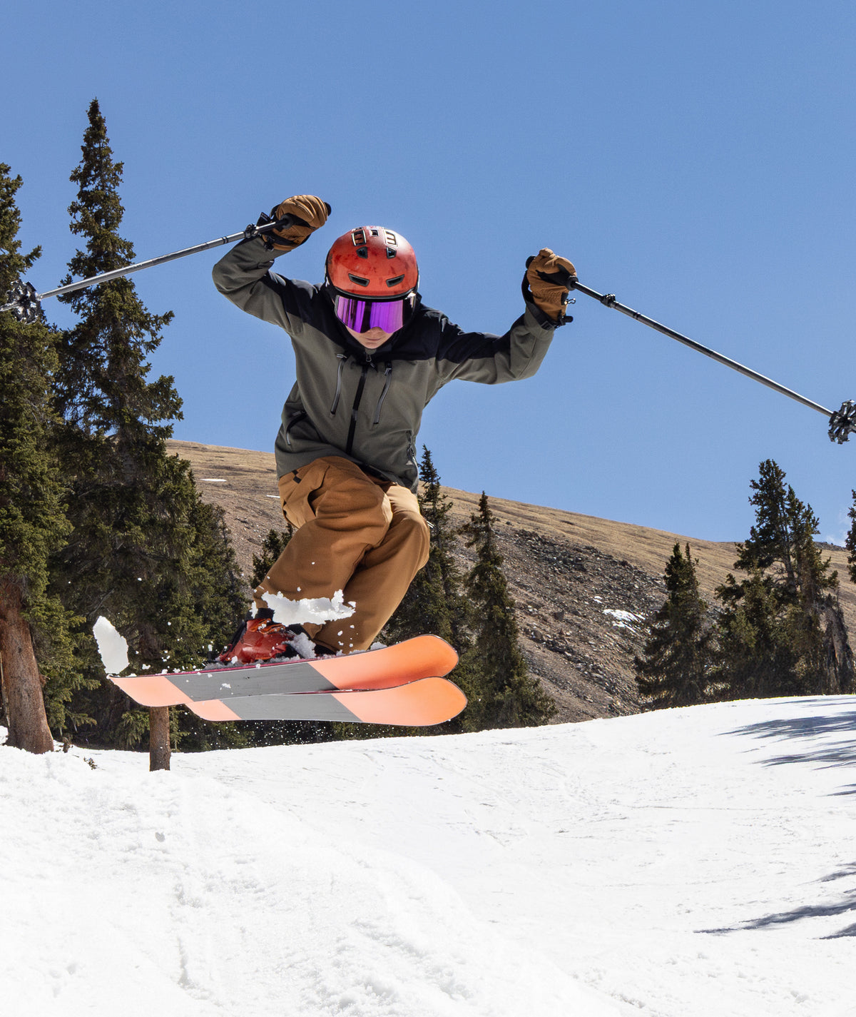 young person skiing on a snowy mountain shown performing a jump wearing a olive green jacket and rust colored pants.