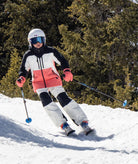 young person shown skiing down a snowy trail surrounded by pine trees shown wearing a black, white, and coral jacket with white pants.