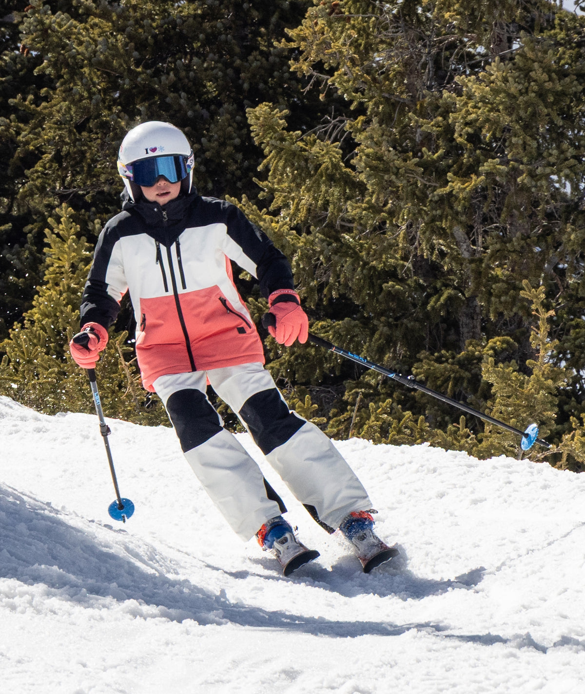 young person shown skiing down a snowy trail surrounded by pine trees shown wearing a black, white, and coral jacket with white pants.