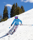 girl shown skiing down a snowy mountain in a light blue jacket with abstract multi color print pants. a blue sky and pine trees can be seen in the background.