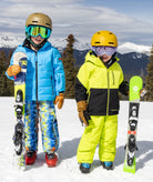 Two children in ski gear standing on a snowy slope with mountains in the background.
