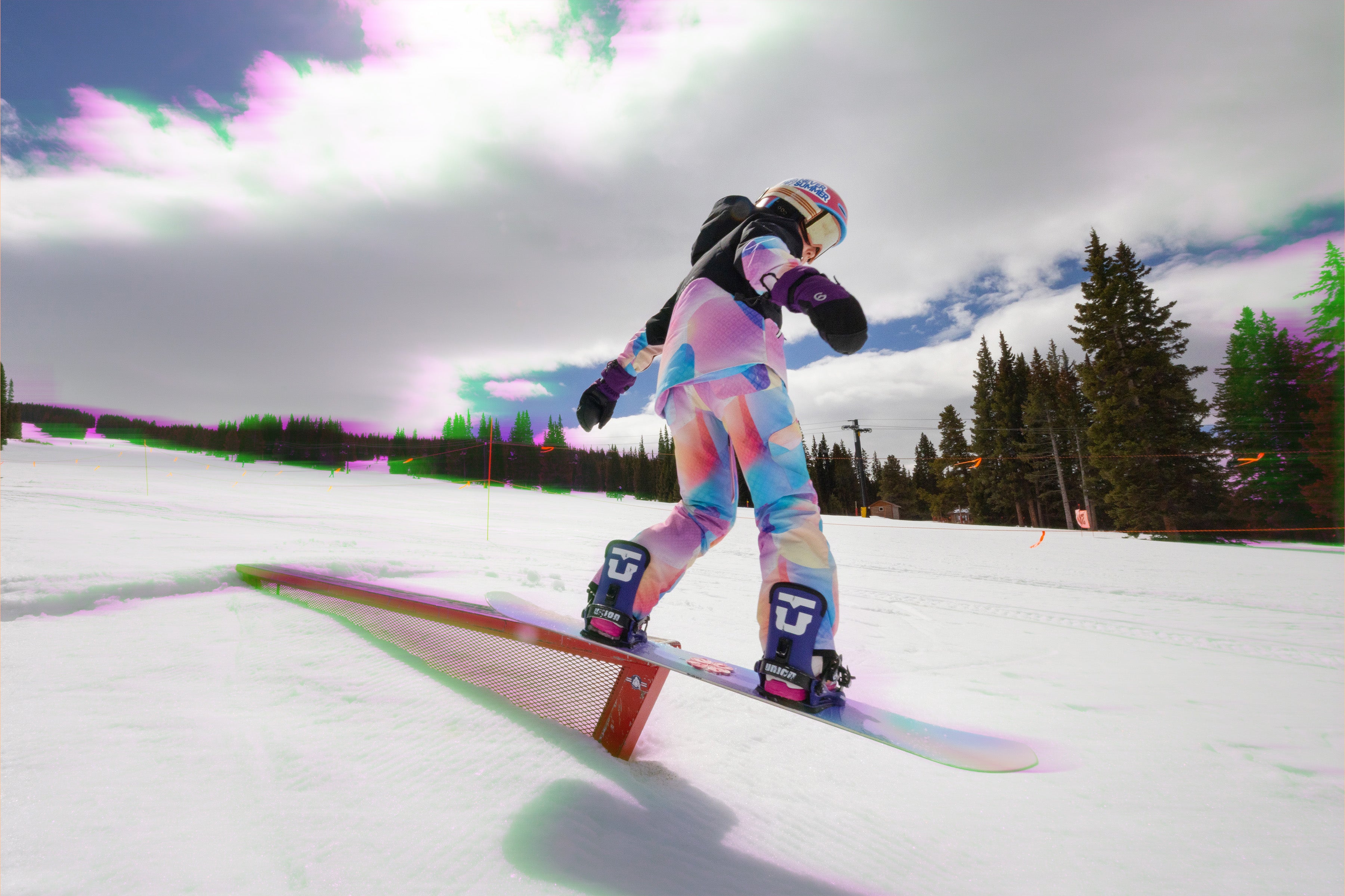 Youth girl snowboarding on a rail with a colorful outfit and helmet against a snowy background.