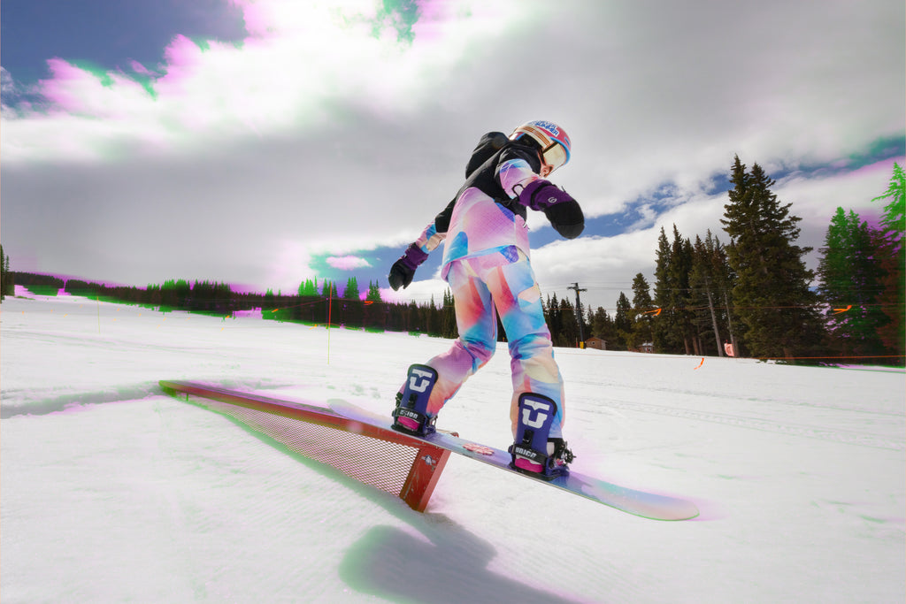 Youth girl snowboarding on a rail with a colorful outfit and helmet against a snowy background.