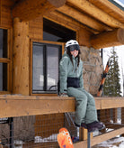 Person in ski gear sitting on a wooden deck with a log cabin in the background