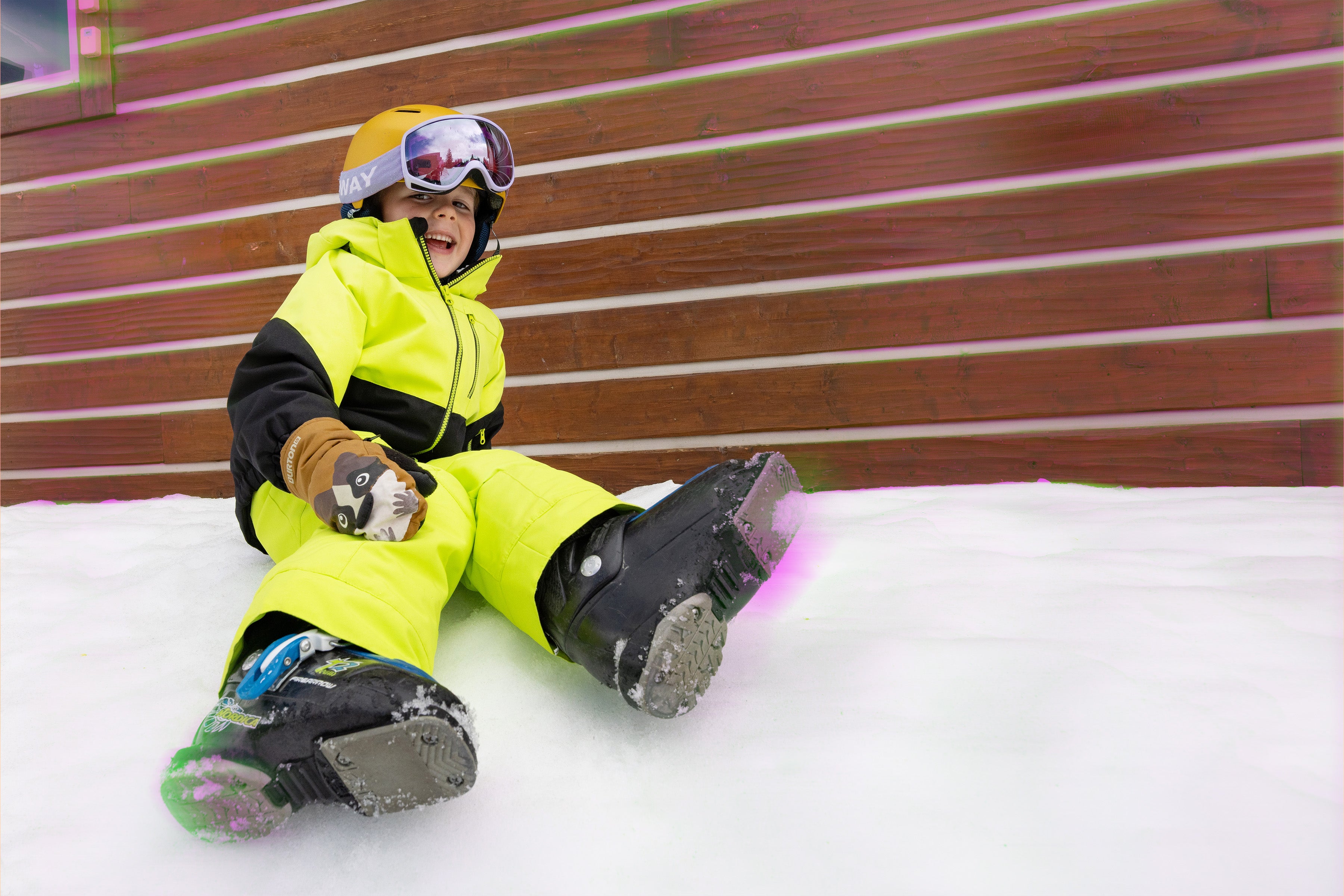 Child in bright yellow snowsuit sitting on a snowy surface with a wooden wall in the background