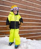 Child in bright yellow and black snow gear standing in the snow against a wooden wall.