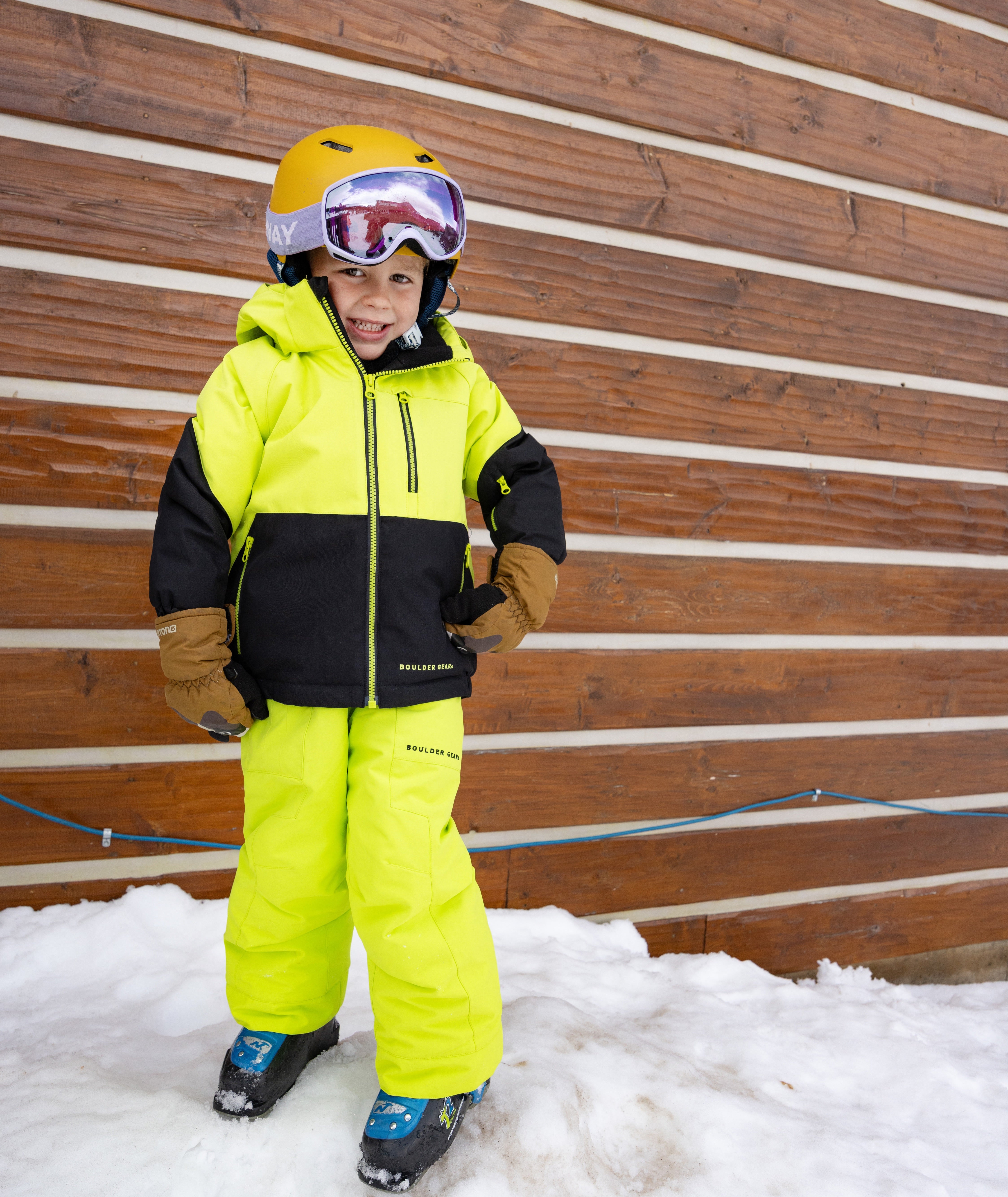 Child in bright yellow and black snow gear standing in the snow against a wooden wall.
