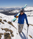 Person carrying skis up a snowy mountain with a scenic view of snow-covered mountains in the background.