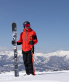 Person in red jacket and black pants holding skis and poles on a snowy mountain with clear blue sky