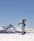 Person in a patterned snowsuit holding skis and poles with mountains in the background